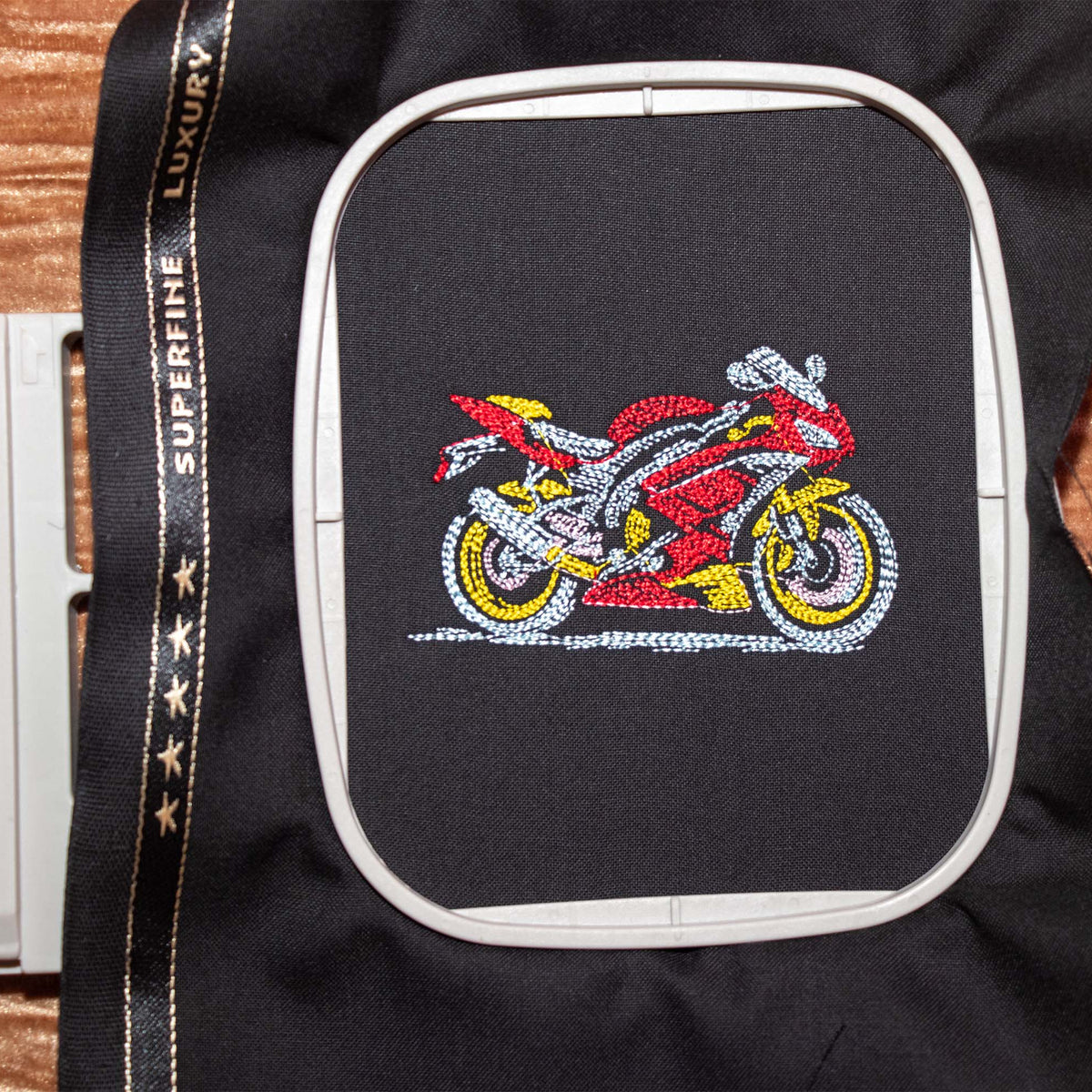 A close-up of a black garment or fabric panel with a white hoop holding a colorful embroidered motorcycle design in red, yellow, white, and gray. A black branded trim strip with gold text is visible along the left side, placed on a brown wood surface.